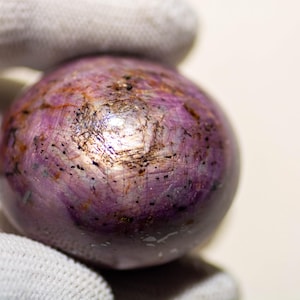 Könnte beinhalten: Nahaufnahme eines polierten, kugelförmigen Edelsteins. Der Stein ist hauptsächlich tiefviolett mit helleren rosa und braunen Streifen. Die Oberfläche hat ein leicht strukturiertes Aussehen mit kleinen dunklen Einschlüssen.