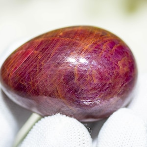 Puede incluir: Una gema pulida en forma de huevo, de un profundo color burdeos, con vetas rojas y anaranjadas más claras. La piedra se sostiene en una mano enguantada de blanco, resaltando su superficie lisa y reflectante. El color de la gema sugiere que podría ser un rubí.