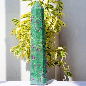 Può includere: Un'alta torre di cristallo a sei facce caratterizzata da un colore di base verde vibrante con numerose inclusioni color magenta incastonate in tutta la pietra. L'esemplare minerale levigato presenta un apice appuntito e una base esagonale.