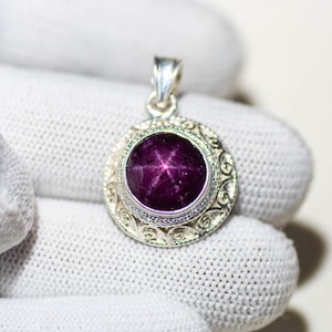 May include: A silver pendant with a large, round, deep purple gemstone. The gemstone has a star-like pattern. The pendant is held by a gloved hand. The pendant has an ornate, circular silver setting.