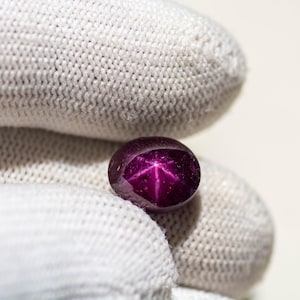 Puede incluir: Una gema pulida, de forma ovalada, de color rojo intenso, con un patrón de estrella de seis rayos. La gema está sujeta por una mano enguantada, mostrando su color y el efecto de estrella interno. Probablemente sea un rubí estrella.