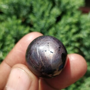 Puede incluir: Una gema esférica oscura con una superficie pulida. La piedra tiene un tono púrpura oscuro con vetas reflectantes, sostenida en una mano contra un fondo verde borroso. La gema parece ser una esfera pulida.
