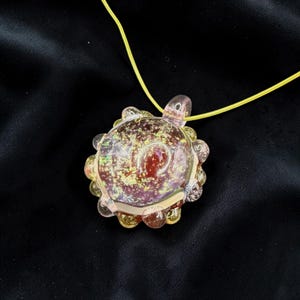 May include: A handcrafted glass turtle pendant with a yellow cord. The pendant features a clear, iridescent body with a reddish-brown center and gold glitter accents. The turtle's shell has a scalloped edge.
