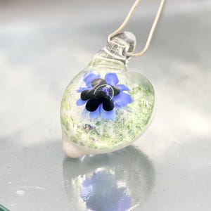 May include: A heart-shaped glass pendant suspended on a silver-toned chain. The clear glass features an embedded blue and black flower design with green speckled accents throughout the interior, creating a layered and dimensional appearance.