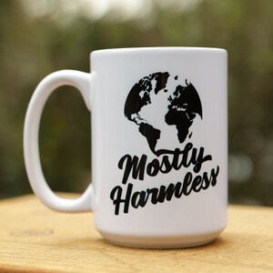 May include: White ceramic mug with a black graphic of the Earth and the words "Mostly Harmless" in a script font. The mug has a large handle and is sitting on a wooden surface.