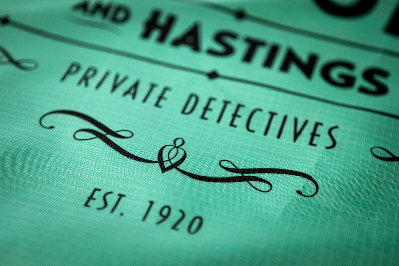 May include: A teal green sign with black text that reads "AND HASTINGS PRIVATE DETECTIVES EST. 1920". The sign has a decorative black flourish with a heart shape in the center.