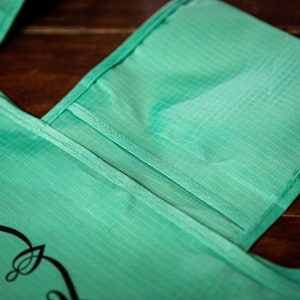 May include: A teal reusable shopping bag with a small pocket on the front. The bag has a black design on the front that says "Shop Local".