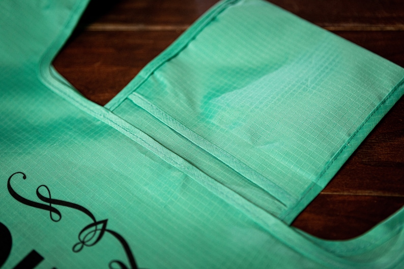 May include: A teal reusable shopping bag with a small pocket on the front. The bag has a black design on the front that says "Shop Local".