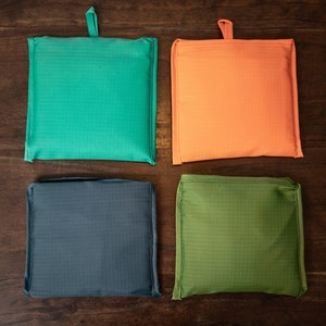 May include: Four foldable shopping bags in different colors: teal, orange, dark blue, and green. The bags are made of a lightweight, water-resistant material and have a simple design.