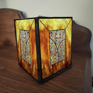 May include: A stained glass lamp with a rectangular shape. The lamp features a warm color palette of amber, orange, and yellow. The design includes a central panel with a terrazzo-like pattern and a stylized tree motif.