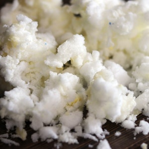 May include: Close-up of a pile of white foam pieces, possibly from a cushion or stuffing. The foam has a textured, irregular surface with some yellow and blue specks visible. The background is a dark brown wooden surface.