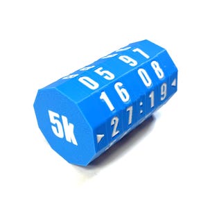 Op de afbeelding: Een blauwe, veelzijdige dobbelsteen met witte cijfers en de tekst "5k". De dobbelsteen heeft verschillende getallen en tijdnotaties, waaronder "05 97", "16 08" en "27:19". Het object staat tegen een witte achtergrond.