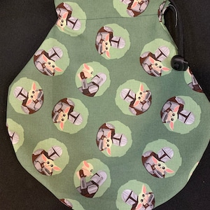 May include: Green drawstring bag with a repeating pattern of a small green alien character holding a small, gray, helmet-shaped object. The alien is wearing a brown, hooded robe.