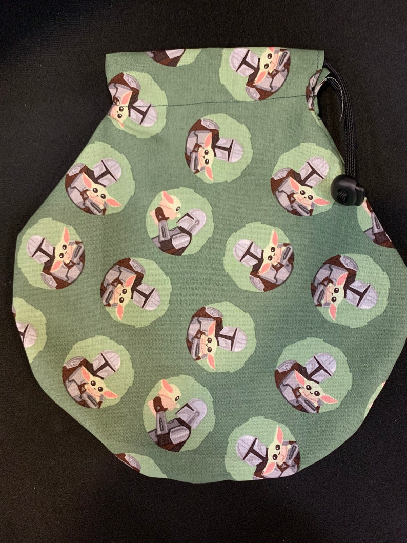 May include: Green drawstring bag with a repeating pattern of a small green alien character holding a small, gray, helmet-shaped object. The alien is wearing a brown, hooded robe.