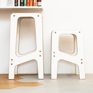 May include: Two white wooden stools with a simple, modern design. The taller stool is slightly behind the shorter stool. Both stools have a rectangular shape with rounded edges.