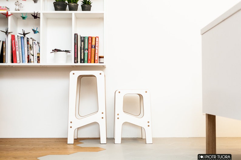 May include: Two white wooden stools with a simple, modern design. The taller stool is slightly behind the shorter stool. Both stools have a rectangular shape with rounded edges.