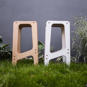 May include: Two wooden stools, one natural wood colour and one white, with a simple, modern design. The stools are standing on a grassy surface.