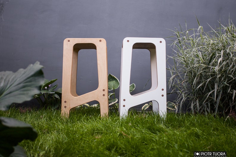 May include: Two wooden stools, one natural wood colour and one white, with a simple, modern design. The stools are standing on a grassy surface.