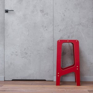 May include: A red wooden step stool with a simple design. The stool is standing on a wooden floor in front of a grey concrete wall.