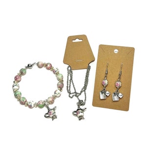 May include: A set of tea-themed jewelry featuring a bracelet, necklace, and earrings. The bracelet and necklace have a teapot charm and are made with pink and green floral patterned beads. The earrings have a floral design and a tea bag charm with the word "tea" on it.