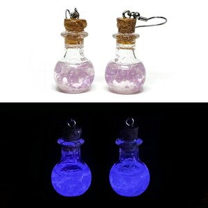 May include: A pair of earrings featuring miniature clear glass bottles with cork stoppers. The bottles are filled with small, light purple spherical beads. In low light, the contents of the bottles emit a bright, glowing blue light.