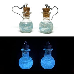 May include: A pair of drop earrings featuring miniature clear glass bottles with cork stoppers. The bottles are filled with a light blue liquid containing small white particles. In low light, the liquid emits a bright blue glow.