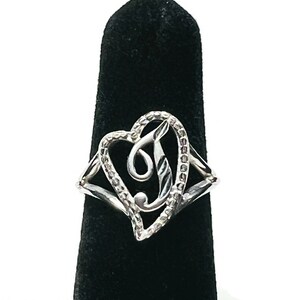 May include: A silver-toned ring featuring a heart-shaped frame with a textured, sparkling surface. Inside the heart, a stylized, cursive letter J is centered. The band of the ring splits into two thin, polished silver-toned arms on each side.