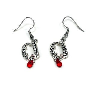 May include: Silver-toned vampire fang earrings with a red teardrop-shaped bead. The earrings feature a detailed fang design and a hook closure. These earrings are a unique accessory for Halloween or costume parties.