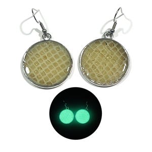 May include: A pair of round drop earrings featuring a textured, cream-colored snakeskin-like pattern encased in silver-toned metal frames with hook backings. Below, the same earrings are shown glowing with a bright green light in the dark.