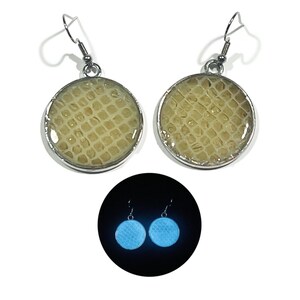 May include: A pair of round drop earrings featuring a textured, cream-colored snakeskin-like pattern encased in silver-toned metal frames with hook wires. A secondary circular inset shows the same earrings glowing with a bright blue light in the dark.