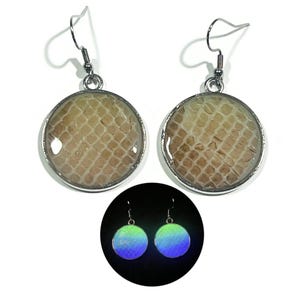 May include: A pair of round drop earrings featuring a beige snakeskin-like texture encased in a silver-toned metal frame with hook wires. A circular inset shows the same earrings glowing with a vibrant blue and green light in the dark.