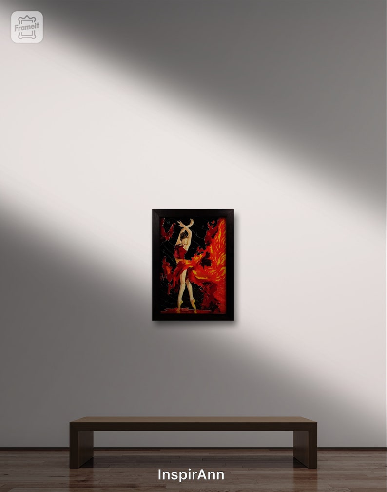 May include: A framed painting of a woman in a red dress dancing with flames behind her. The painting is on a wooden bench in front of a white wall.