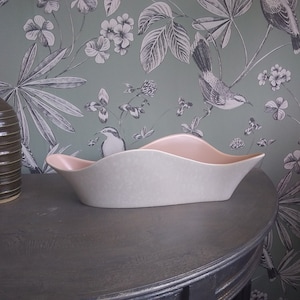 Può includere: Un vaso da fiori in ceramica bianca con un bordo rosa. Il vaso ha una forma ondulata e organica ed è appoggiato su un tavolo di legno scuro.