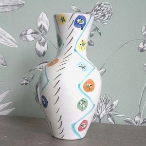 May include: White ceramic vase with a colorful floral design. The vase features a slender neck and a wider base, adorned with blue, orange, green, and yellow flower-like patterns and a turquoise zig-zag pattern. Black diagonal lines add further detail.