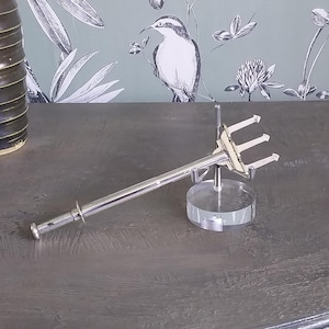 May include: A silver serving fork with three prongs, presented on a clear acrylic stand. The fork has a long handle and a decorative top. The background features a grey surface and wallpaper with a bird and floral pattern.