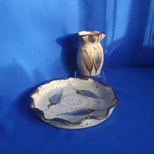 May include: A set of two handmade pottery pieces, a small vase and a decorative plate. Both pieces feature a white glaze with brown and blue accents and a wavy edge.