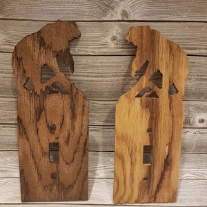 Puede incluir: Dos cubiertas de interruptor de luz de madera con un diseño de silueta de oso. Los osos se enfrentan y están de pie sobre un tocón de árbol. La madera es de color marrón oscuro con un patrón de veta de madera.