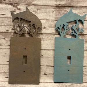 May include: Two wooden light switch covers. One is stained a dark brown, the other is painted a light blue. Each features a dolphin leaping above coral. The switch opening is rectangular.