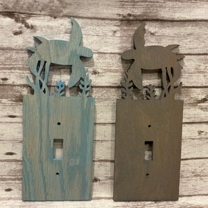 May include: Two wooden light switch covers with a cutout design of a sea turtle and seaweed. The cover on the left is painted a light blue-gray color, while the cover on the right is a dark brown color.