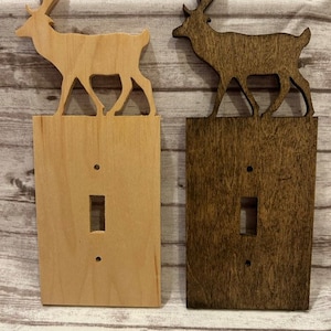 Könnte beinhalten: Zwei Holz-Lichtschalterabdeckungen mit einer Hirsch-Silhouette auf der Oberseite. Die Abdeckung links ist aus Naturholz und die Abdeckung rechts ist dunkelbraun gebeizt.