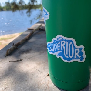 May include: Green insulated travel mug with a blue and white sticker that says "Lake Superior" with a graphic of the lake outline.