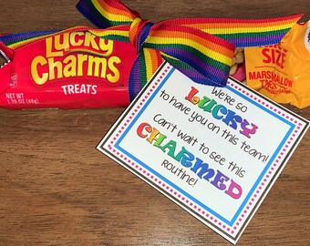 May include: A white card with a rainbow border and the text "We're so lucky to have you on this team! Can't wait to see this charmed routine!"  The card is tied with a rainbow ribbon to a package of Lucky Charms cereal and a bag of marshmallows.