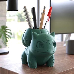 Puede incluir: Un organizador de escritorio de color verde oscuro con acabado mate, con forma de criatura y una estructura vegetal bulbosa en la espalda. El bulbo hueco sirve como recipiente para cuatro bolígrafos, incluyendo instrumentos de escritura plateados, negros, de madera y con tapa roja.