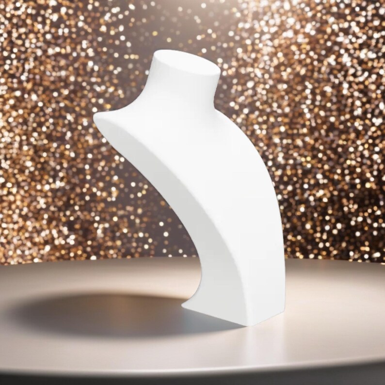 May include: A white jewelry display bust, designed to showcase necklaces. The bust has a curved, elegant shape, with a smooth, matte finish. The background is a blurred display of gold glitter.