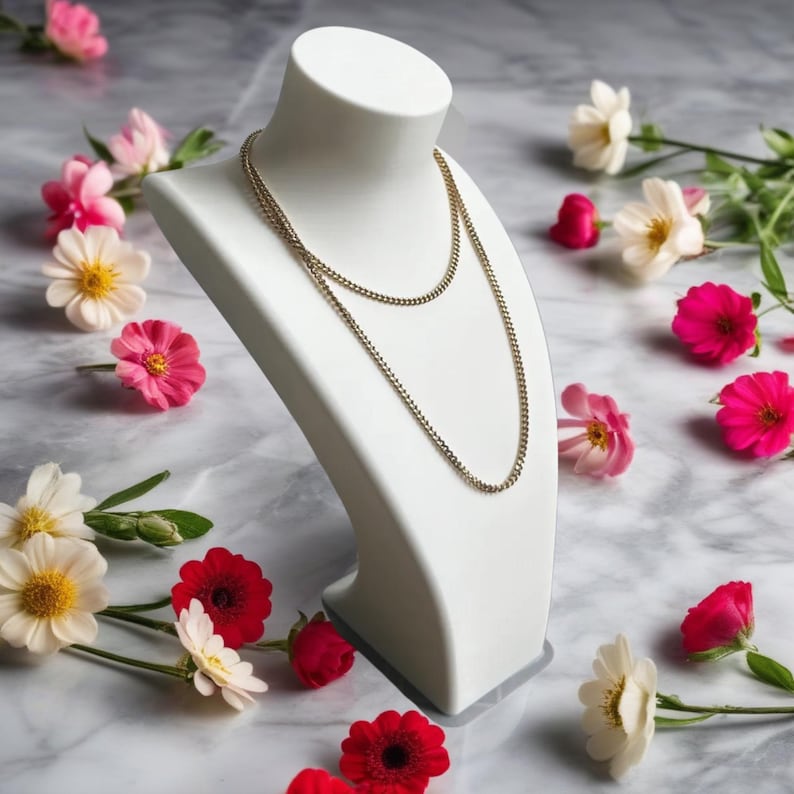 May include: A gold chain necklace displayed on a white jewelry bust. The necklace is draped in two strands. The bust is surrounded by pink, red, and white flowers, set against a marble background. The jewelry is a classic accessory.