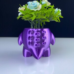 Pu&ograve; includere: Un vaso viola, stampato in 3D, a forma di creatura, con fiori artificiali. Il vaso ha una superficie strutturata con motivi decorativi. I fiori sono bianchi e azzurri, con foglie verdi.