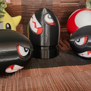 May include: A collection of black, 3D-printed Bullet Bill figures with red and white facial details. The figures are arranged on a textured surface, with a yellow star-shaped figure and a red and white ball in the background.