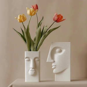 May include: Two white, textured face vases, one rectangular and one profile, with a bouquet of tulips in the rectangular vase. The tulips are yellow and orange, with green stems and leaves. The vases are on a neutral surface.