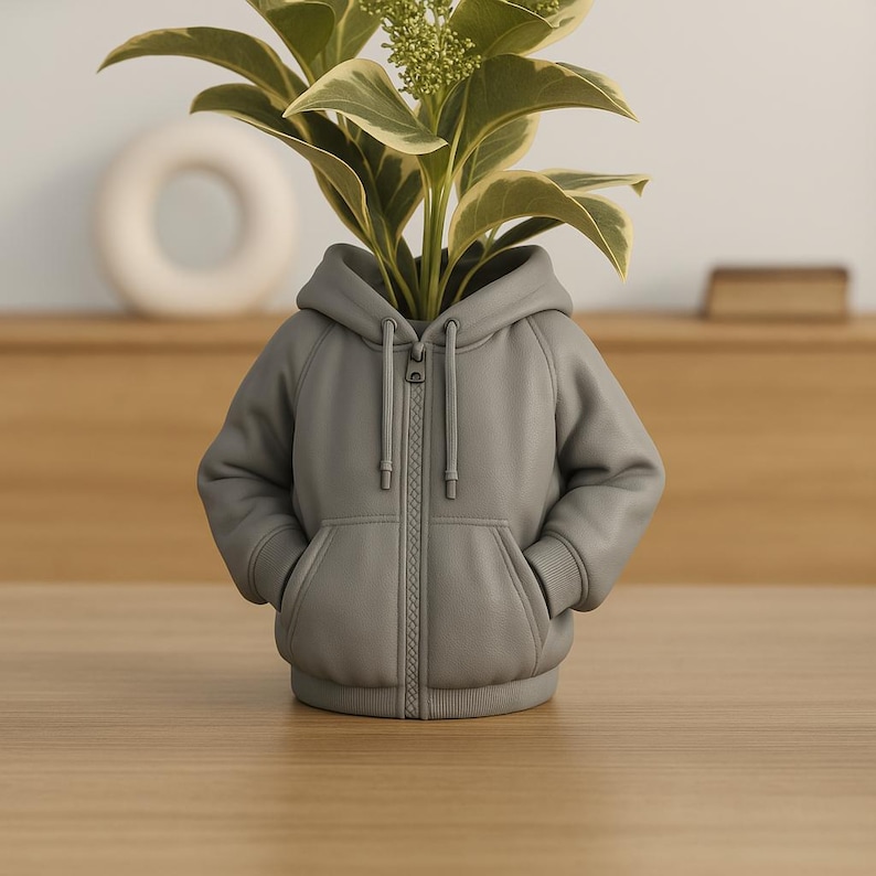 Peut inclure: Un pot de fleurs en forme de sweat &agrave; capuche gris avec une fermeture &eacute;clair compl&egrave;te et des poches avant, contenant une plante aux feuilles vertes et cr&egrave;me. Le pot est pos&eacute; sur une surface en bois.
