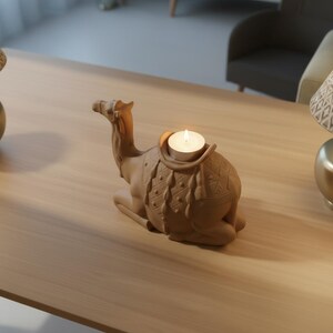 May include: A tan-colored camel-shaped candle holder with a lit tea light candle. The decorative piece has detailed saddle designs and is placed on a light-colored wooden surface. The candle holder is a unique home decor item.
